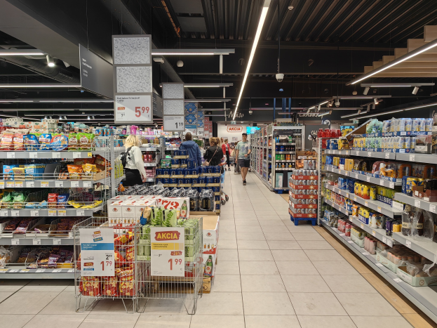 Innensicht eines Supermarkts mit Einkaufenden, Lebensmittelregalen, beschrifteten Einkaufswagen, Informationsschildern, Deckenleuchten und sichtbarem Boden.