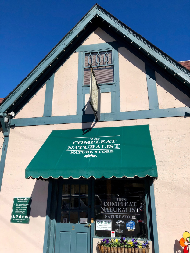 Ein Gebäude mit einem grünen Vordach, einer Tür, Fenstern, einem Schild mit der Aufschrift "The Complete Naturalist Nature Store" und einem Korb mit blühenden Pflanzen unter einem bewölkten Himmel.