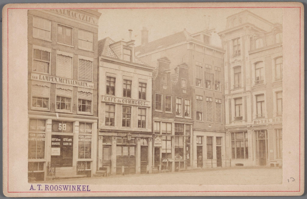 Altes Schwarz-Weiß-Foto einer Stadtstraße mit mehrstöckigen Gebäuden, die Fenster, Türen und Schilder zeigen, mit undeutlicher Schrift am unteren Rand.