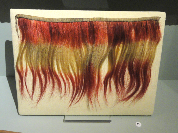 Nahaufnahme einer farbenfrohen Haarsträhne mit roten, gelben und cremefarbenen Strähnen, die auf einem Brett in einer Museumsausstellung präsentiert wird.