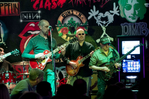 Drei Männer auf einer Bühne, die Gitarren spielen, mit einer Menge unten und einem Graffiti oder einer Wandmalerei im Hintergrund.