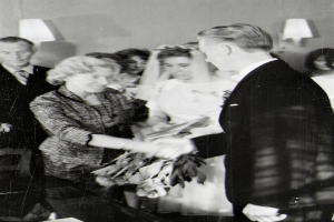 Ein Schwarz-Weiß-Foto eines Brautpaares beim Austausch der Trauringe, wobei die Braut in einem weißen Kleid und Schleier mit einem Strauß Blumen in der Hand zu sehen ist und der Bräutigam in einem Anzug und Krawatte steht, mit Wandlampen im Hintergrund.