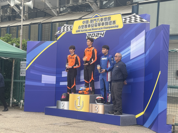 Vier Männer auf einem Podest mit Trophäen in den Händen, eine Person links davon gehend und andere rechts stehend, eine Tafel mit Text, Helme, Gebäudedecke, Pflanzen und ein Zelt im Hintergrund.