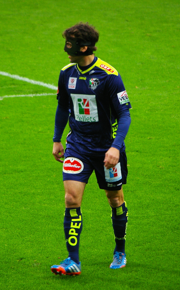 Ein Mann steht auf einem Fußballfeld und trägt ein blaues T-Shirt, Shorts, Socken, Schuhe und eine Gesichtsmaske.