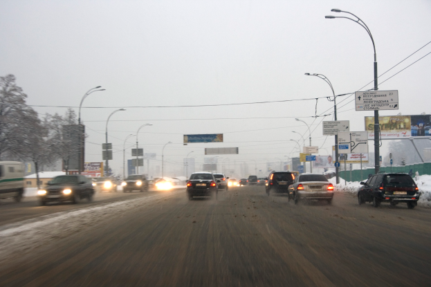 Eine belebte Stadtstraße mit Fahrzeugen auf einer schneebedeckten Straße, gesäumt von Laternenmasten, Texttafeln, Bäumen und Gebäuden unter einem verschneiten Himmel.