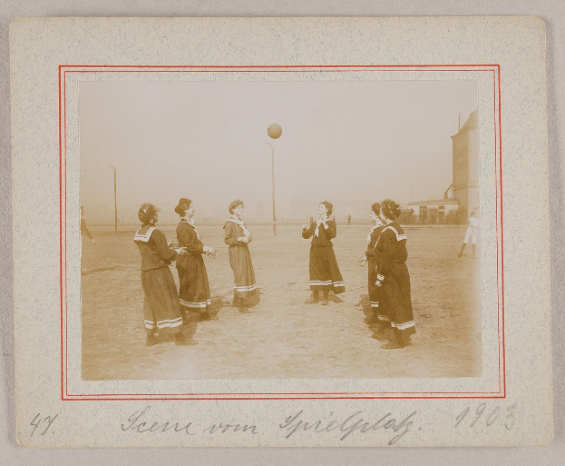 Eine Gruppe von Frauen, die auf dem Boden Fußball spielen, mit einem Ball in der Mitte, Pfosten und Gebäuden im Hintergrund und Text am unteren Bildrand.