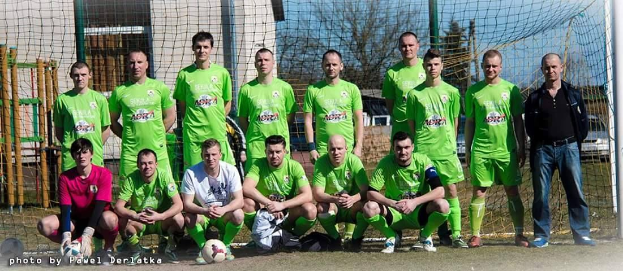 Eine Gruppe von Männern in grünen T-Shirts steht auf einem Fußballfeld, einige in Hockstellung, mit einem Ball und einem Torpfosten im Hintergrund; Gebäude, Bäume und Fahrzeuge sind im Hintergrund zu sehen.