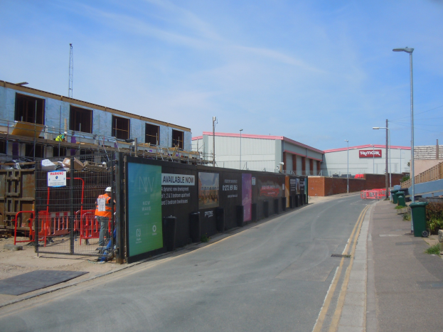 Eine Straße mit einem Gehweg auf der rechten Seite, mit Laternen, Mülltonnen, Pflanzen und einem Zaun, die zu Gebäuden und einem Turm im Hintergrund führt, wobei auf der linken Seite eine Baustelle zu sehen ist.