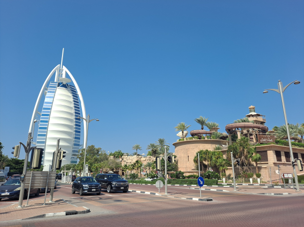 Ikonisches Burj Al Arab Hotel in Dubai, VAE, mit Fahrzeuge, Verkehrszeichen, Schildern, Pflanzen, Bäumen und Gebäuden im Vordergrund und einem sichtbaren Himmel.
