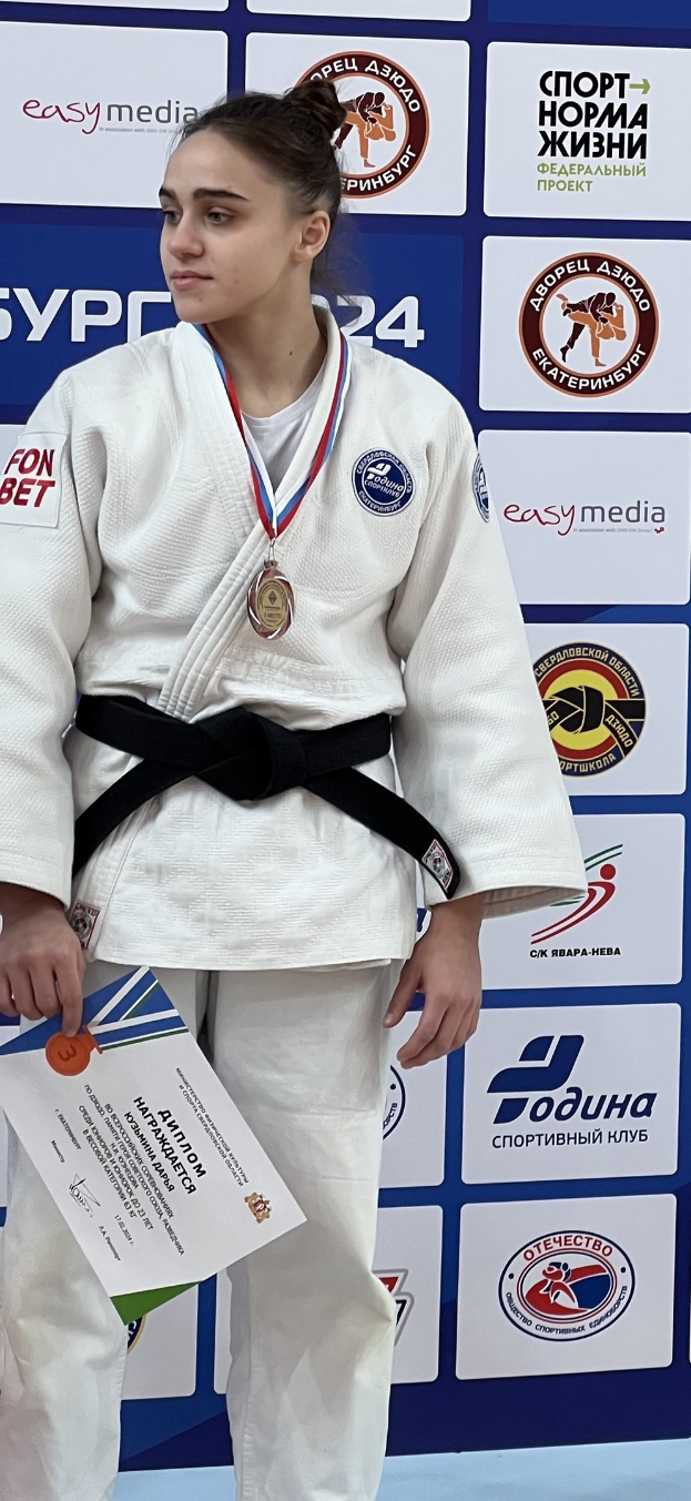 Frau in weißem Judo-Anzug mit schwarzem Gürtel auf einem Podest stehend, eine Medaille und ein Zertifikat haltend, mit einem logo-bedeckten Hintergrundbrett.