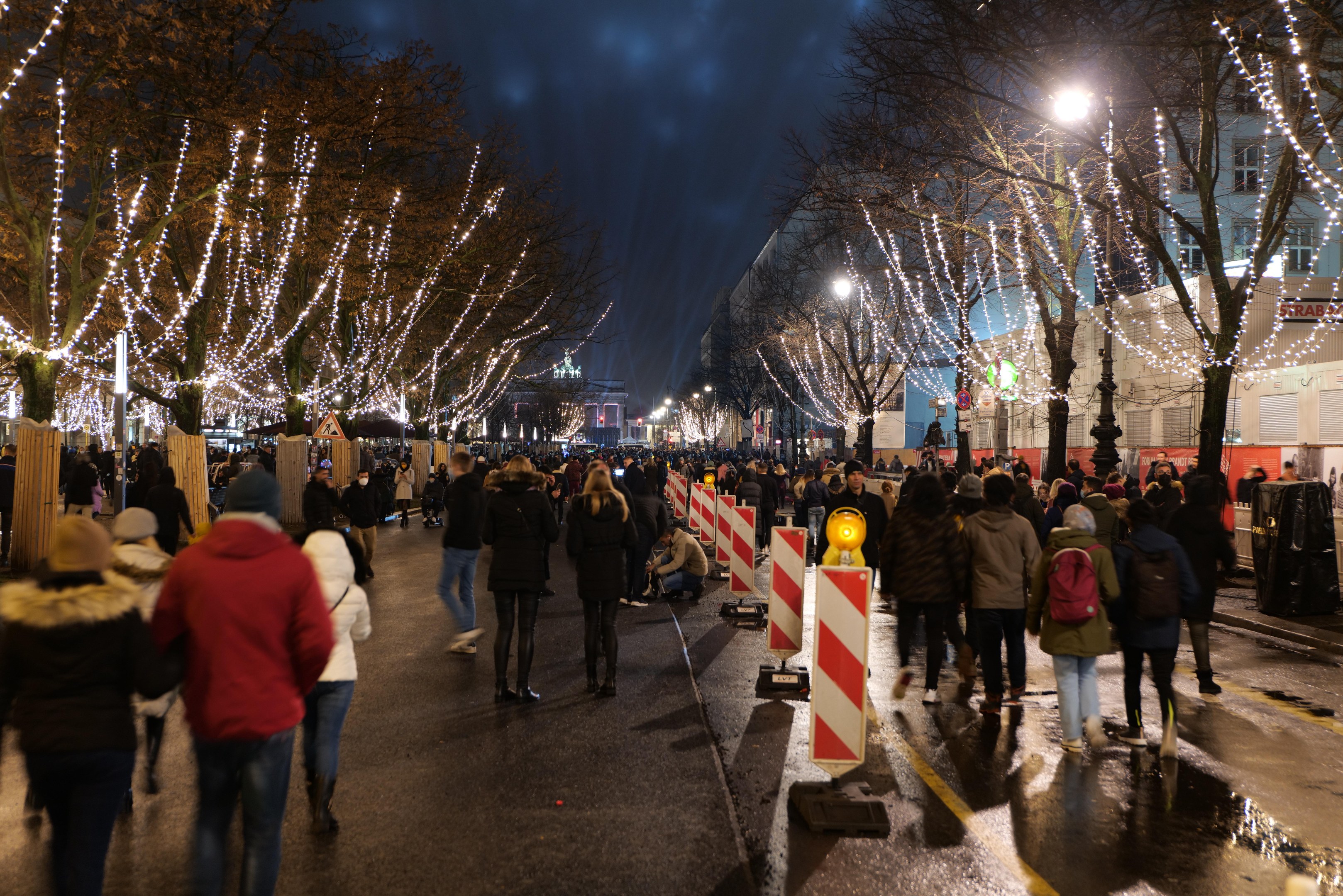 Eine Menschenmenge, die nachts eine Straße entlanggeht, beleuchtet von festlichen Weihnachtslichtern mit Bäumen und Gebäuden im Hintergrund unter einem bewölkten Himmel.