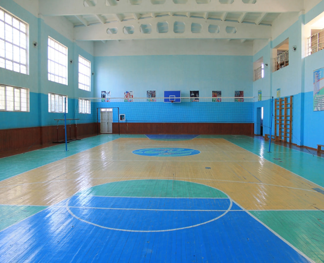 Ein geräumiger Turnsaal mit einem blauen und weißen Boden, der einen Basketballplatz, Netz, Stangen, eine Tür, Fenster, Plakate an den Wänden, ein Geländer und ein Dach zeigt.