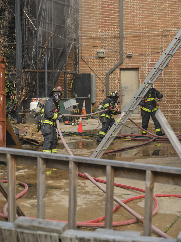 Feuerwehrleute in Helmen arbeiten daran, ein Gebäudefeuer zu löschen, während sie Schläuche halten, mit einem Metallzaun, Rohren, einem Behälter, einem Verkehrskegel, Gegenständen, Fenstern, einer Tür, einem Metallrahmen, Drähten, einem Baum und Himmel.