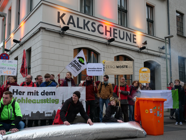 Eine Gruppe von Menschen mit Protestschildern und Transparenten vor einem Gebäude in Deutschland, wobei zwei Personen auf einem Gegenstand im Vordergrund sitzen und ein Müllcontainer rechts zu sehen ist.