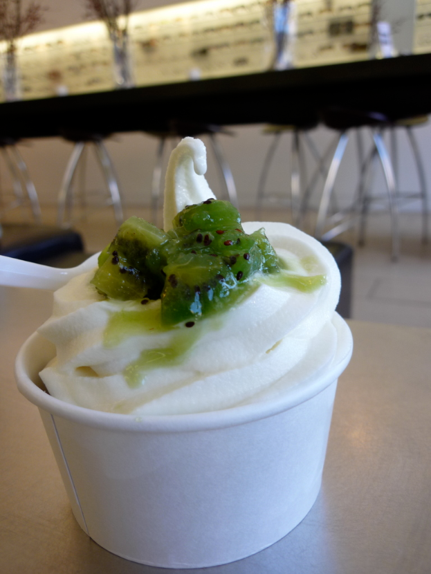 Ein Cupcake auf einem Tisch mit Stühlen und Tischen im Hintergrund und Lampen und Röhrenlampen oben.