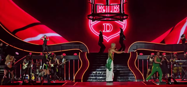 Eine Gruppe von Menschen auf einer Bühne, einige halten Musikinstrumente, mit Stufen, die zur Bühne führen, und einem Schild im Hintergrund mit der Aufschrift "Rihanna - Live in der O2 Arena, London".