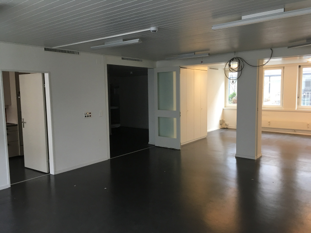 Leerer Büroraum mit schwarzen Böden, weißen Wänden, elektrischen Lampen, Kabeln, Türen, Schränken, Fenstern und einem Blick auf Gebäude und Bäume draußen, verfügbar zur Miete in Amsterdam.