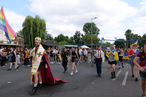 Menschen bei einer Parade mit einer Regenbogenflagge und Musikinstrumenten, mit Laternenmasten, Bäumen und Hütten im Hintergrund.