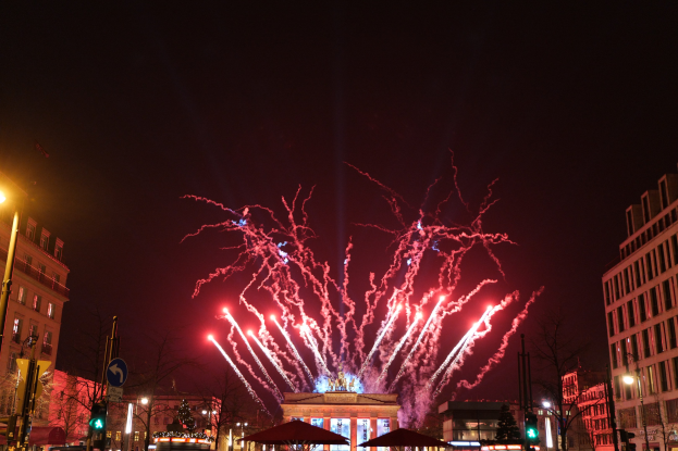 Eine belebte Stadtstraße in Berlin am Silvesterabend, voller Menschen, Fahrzeuge und festlicher Dekoration, mit Gebäuden und Feuerwerk, das den Nachthimmel erhellt.