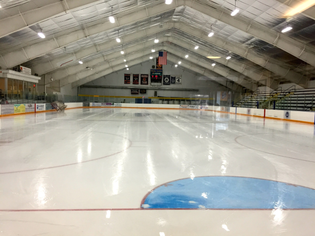 Große Indoor-Eisfläche mit Deckenbeleuchtung, Werbetafeln, Treppen mit Geländern, einer Wand im Hintergrund und einigen Menschen, die auf eine Sportanlage hinweisen, die likely für Hockey verwendet wird.