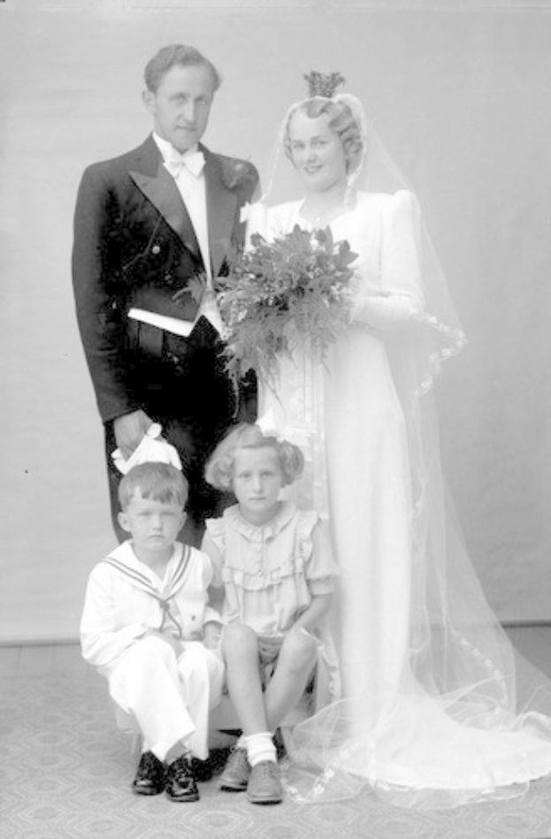 Schwarzes und weißes Foto einer Braut in einem weißen Kleid mit einem Strauß, ein grinsender Bräutigam neben ihr und zwei Kinder, die vor ihnen auf dem Boden sitzen.