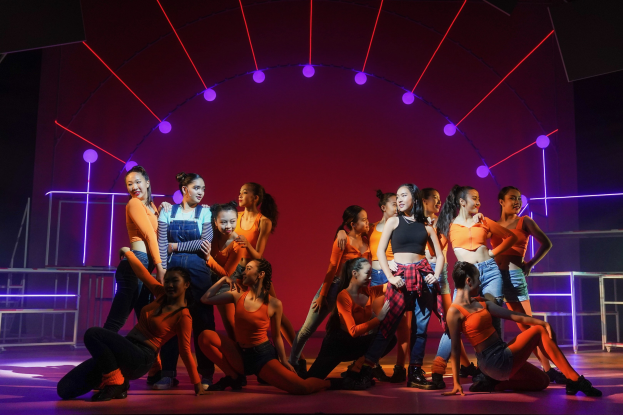 Eine Gruppe junger Tänzer in bunten Kostümen performs auf einer Bühne mit Geländern, ihre Arme erhoben, während sie von bunten Lichtern und Mustern umgeben sind und eine applaudierende Menge zusieht.