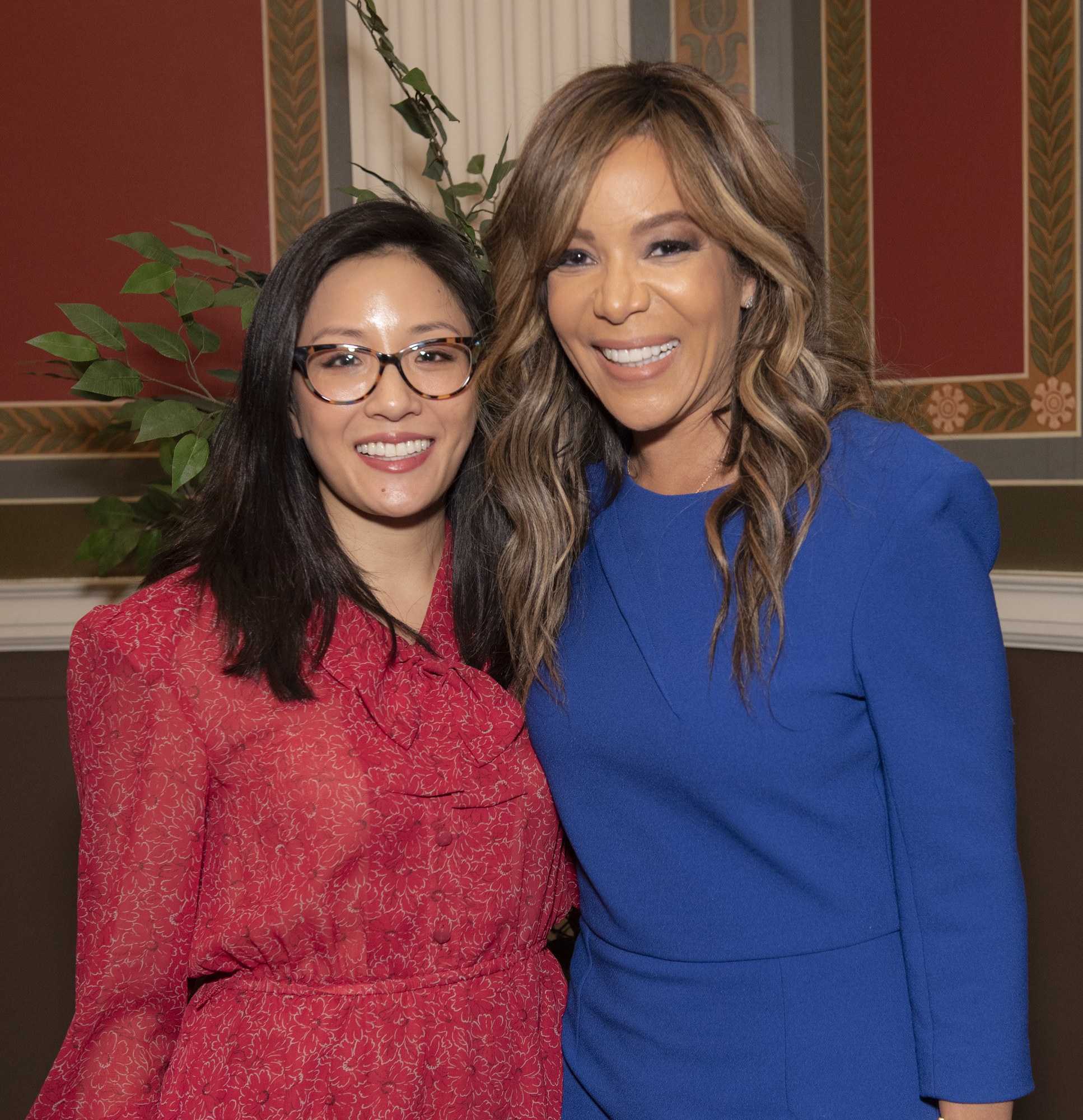Zwei Frauen, die zusammen auf einem Event lächeln, eine in einem blauen Kleid und die andere in einem roten Kleid mit Brille, vor einem Hintergrund aus Pflanzen und Vorhängen.