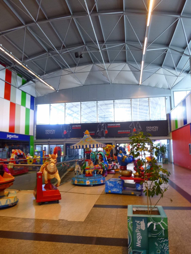 Gro├čer Indoorraum in einem Einkaufszentrum mit verschiedenen Vergnügungsger├Ąten, einem Topfpflanze, verstreuten Spielzeugen, Informationsschildern, Deckenleuchten und Metalltr├Ągern.