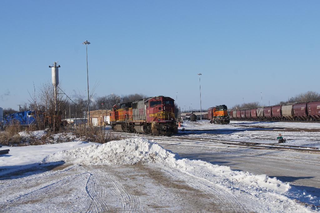 Eine Lokomotive ist vorne positioniert mit Eisenbahnwagen auf der rechten Seite, geparkt auf Schienen mit Schnee auf dem Boden; trockene Bäume und ein Strommast sind im Hintergrund sichtbar.