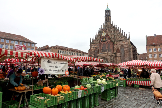 Ein belebter Markt in Nürnberg, Deutschland, mit verschiedenen Früchten und Gemüsen auf dem Stand, Einkaufenden, Zelten und Gebäuden mit einem Uhrenturm im Hintergrund unter einem klaren Himmel.