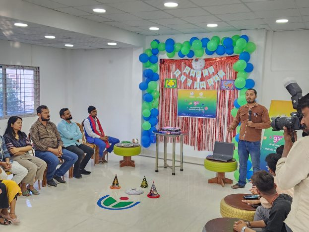 Eine Gruppe von Menschen, die auf Stühlen vor einem Publikum sitzen, mit einer Person, die ein Mikrofon hält, einem Laptop auf einem Hocker, einem Tisch mit verschiedenen Gegenständen, einer Fahne mit Text, Luftballons, einem Vorhang, einem Fenster und einer Decke mit Lampen, die einen 9. Geburtstag feiern.