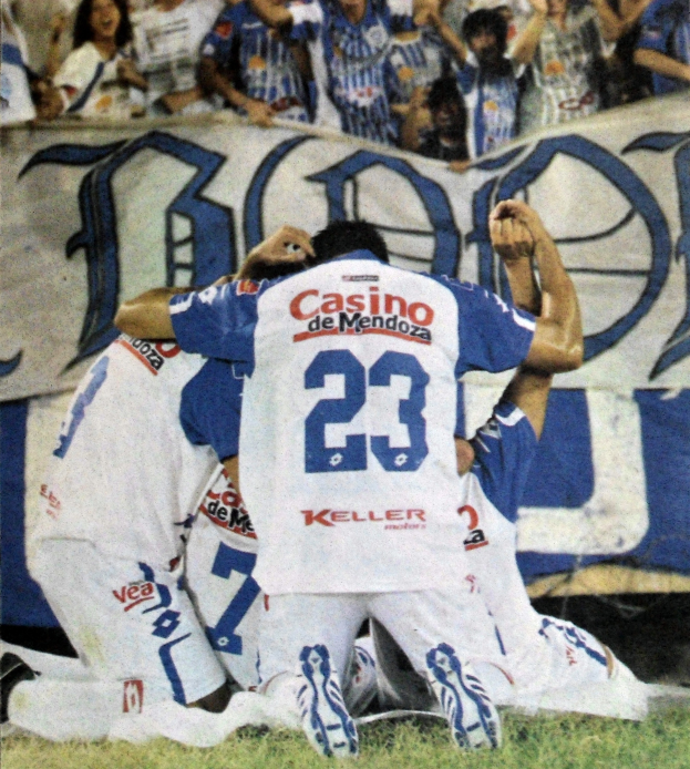 Eine Gruppe von Fussballspielern feiert ein Tor auf dem Platz vor einer jubelnden Menge, mit einem Banner mit Text im Hintergrund.