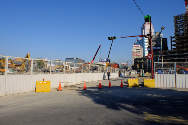 Baustelle mit Verkehrskegeln, eine Person zwischen Absperrungen und einem Zaun, ein Schild, Straßenlaternen, eine Ampel, Gebäude mit Fenstern, Kräne, Bäume und ein bewölkter Himmel.