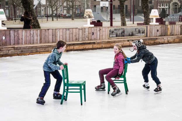 Kinder beim Skifahren vor einem Spielplatz, mit drei Kindern und zwei StÃ¼hlen in der Mitte und GebÃ¤uden, BÃ¤umen, BÃ¤nken, PfÃ¤hlen und einem Basketballfeld im Hintergrund.