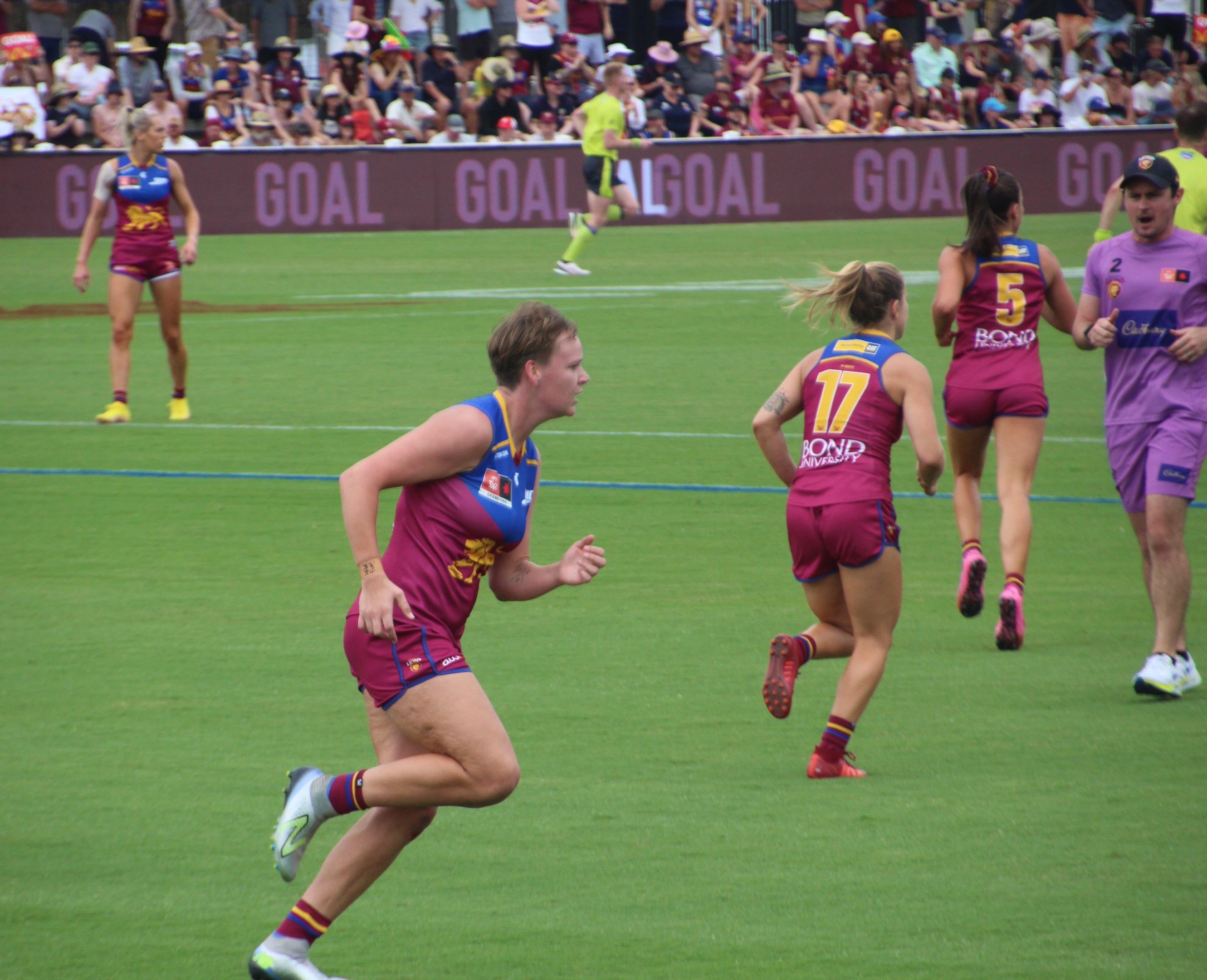 Frauen spielen Australian Rules Football auf einem grünen Feld mit Zuschauern im Hintergrund.