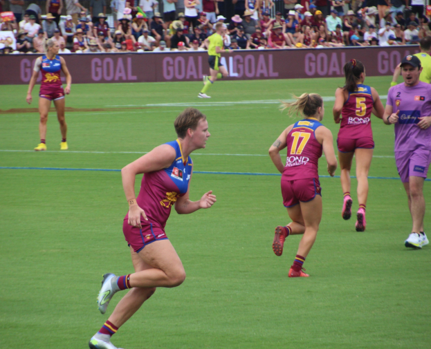 Frauen spielen Australian Rules Football auf einem grünen Feld mit Zuschauern im Hintergrund.