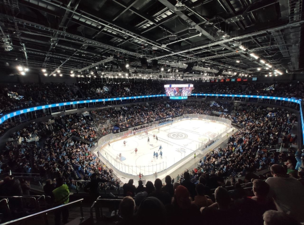 Hockey-Spiel in einer großen Arena mit Zuschauern auf den Tribünen und Mitarbeitern am Boden, mit Geländern, Überwachungsschirmen und heller Beleuchtung, als O2 Arena in London identifiziert.