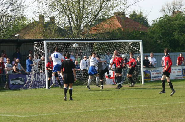 Spieler spielen FÃ¼ßball auf einem Feld mit einem Tor, wÃ¤hrend Zuschauer dahinter stehen, mit BÃ¤umen und HÃ¤usern im Hintergrund.