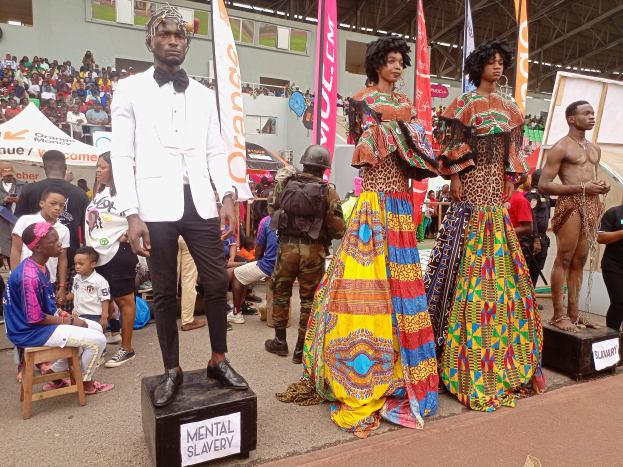 Eine Gruppe von Menschen auf einer Bühne mit einer Schaufensterpuppe in einem weißen Anzug in der Mitte, andere sitzen auf Hockern links und eine Person hält eine Kamera rechts, mit Bannern, Flaggen und einem Gebäude im Hintergrund bei einer Fashion Show in Nigeria.