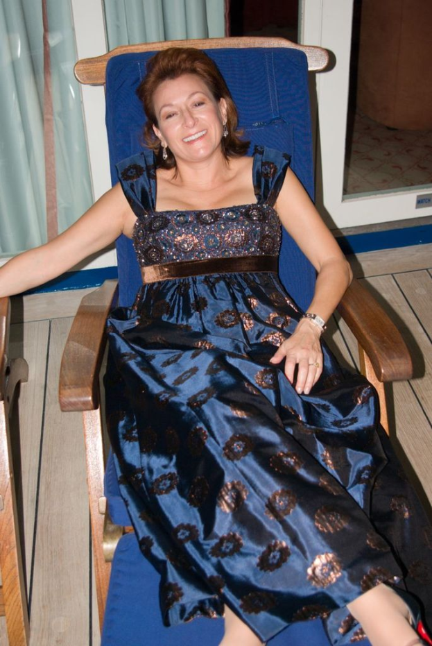 Eine Frau in einem blauen Kleid sitzt auf einem Stuhl und raucht, mit Vorhängen und Glastüren im Hintergrund.