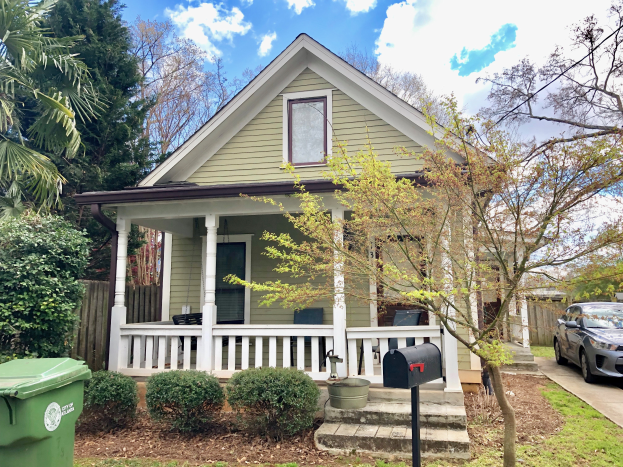 Ein grünes Haus mit einem vor dem Haus geparkten Auto, umgeben von Bäumen, Pflanzen und Gras, mit einem Müllcontainer in der Nähe, mit Fenstern, Säulen, Geländern und einem Briefkasten mit Briefschlitz, unter einem bewölkten Himmel in Charleston, SC 29401.