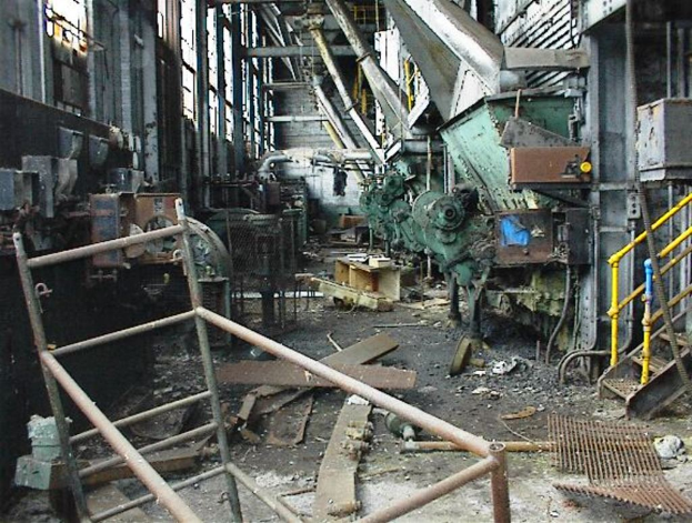 Ein altes, unbenutztes Fabrikinnere mit Maschinen, Schutt auf dem Boden und Fenstern, das Geländer auf beiden Seiten zeigt.
