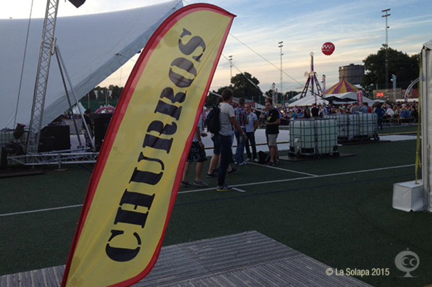 Ein buntes Volksfest-Szenario mit Zelten, einem zentralen Mast, Menschen, Bäumen, Strommasten, Straßenbahnen auf der Straße und einem Eisengestell links.