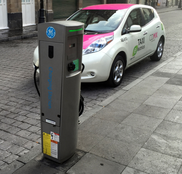Ein Elektroauto steht auf der Straße neben einer Ladestation, im Hintergrund sind Gebäude mit Türen, Läden und einem Pfahl zu sehen.