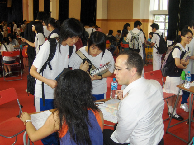 Ein Klassenzimmer mit mehreren M√§dchen in wei√üen T-Shirts, die B√ľcher in der Vornahme halten, ein Mann und eine Frau, die im Vordergrund sitzen und diskutieren, und eine gelbe Wand mit einem Glasfenster im Hintergrund.