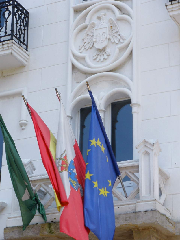 Drei Flaggen, die die Europäische Union darstellen, hängen an der Seite eines Gebäudes mit Fenstern und einem Geländer.