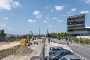 Baustelle mit parkenden Autos, Gebäuden, Straßeninfrastruktur, Vegetation, Sand, Kränen, Bahnschienen, Strommasten, Hügel und einem bewölkten Himmel.