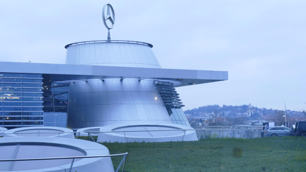 Das Mercedes-Benz Museum in Frankfurt, Deutschland, mit seinem Logo auf dem Dach, umgeben von Gras, Geländern, Fahrzeugen auf der Straße, Bäumen im Hintergrund und einem klaren blauen Himmel.