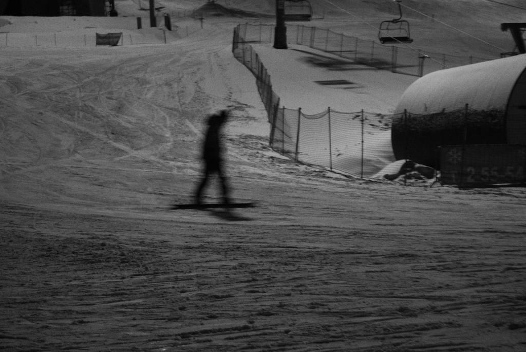 Eine Person, die Snowboard fährt, auf einem schneebedeckten Boden mit einem Zaun und verschiedenen Gegenständen im Hintergrund.