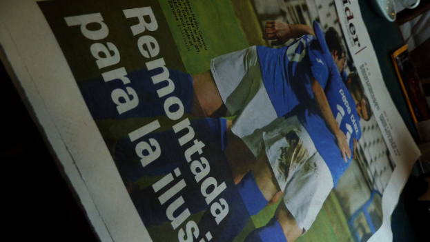 Eine Zeitung und eine Untertasse auf einem Tisch, wobei die Zeitung drei Männer in blauen Hemden zeigt, die möglicherweise Fußball spielen, und Text am unteren Rand des Bildes.